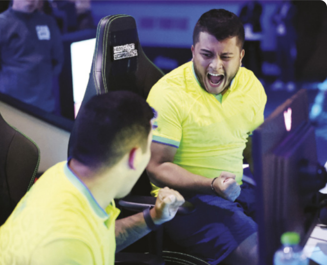 Fotografia de dois homens comemorando com gritos e gestos com os punhos fechados, sentados diante de computadores, uniformizados com camisetas da seleção brasileira.