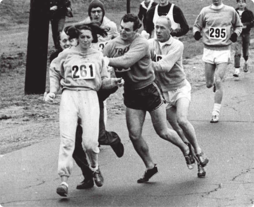 Fotografia de Kathrine Switzer. Ela é uma mulher com roupas de ginástica em uma pista, correndo, com uma placa sobre o peito com o número 261 estampado. Atrás dela estão homens correndo e tentando segurá-la, alguns deles também corredores participantes da corrida.