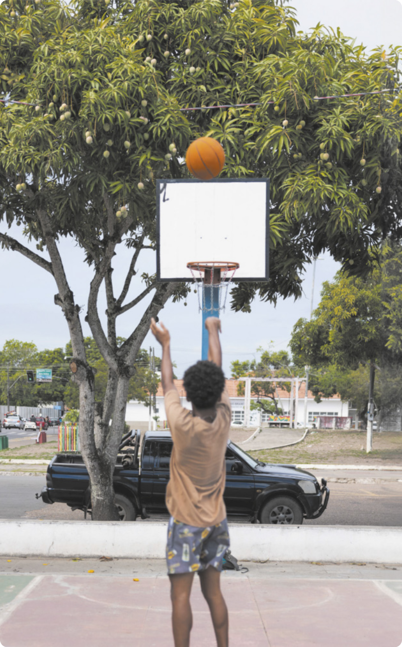 Fotografia de um menino de costas, arremessando uma bola de basquete em direção a uma cesta em uma tabela de basquete, em uma quadra pública em uma praça. A quadra está ao lado de uma via pública com carros estacionados, em uma cidade interiorana arborizada.