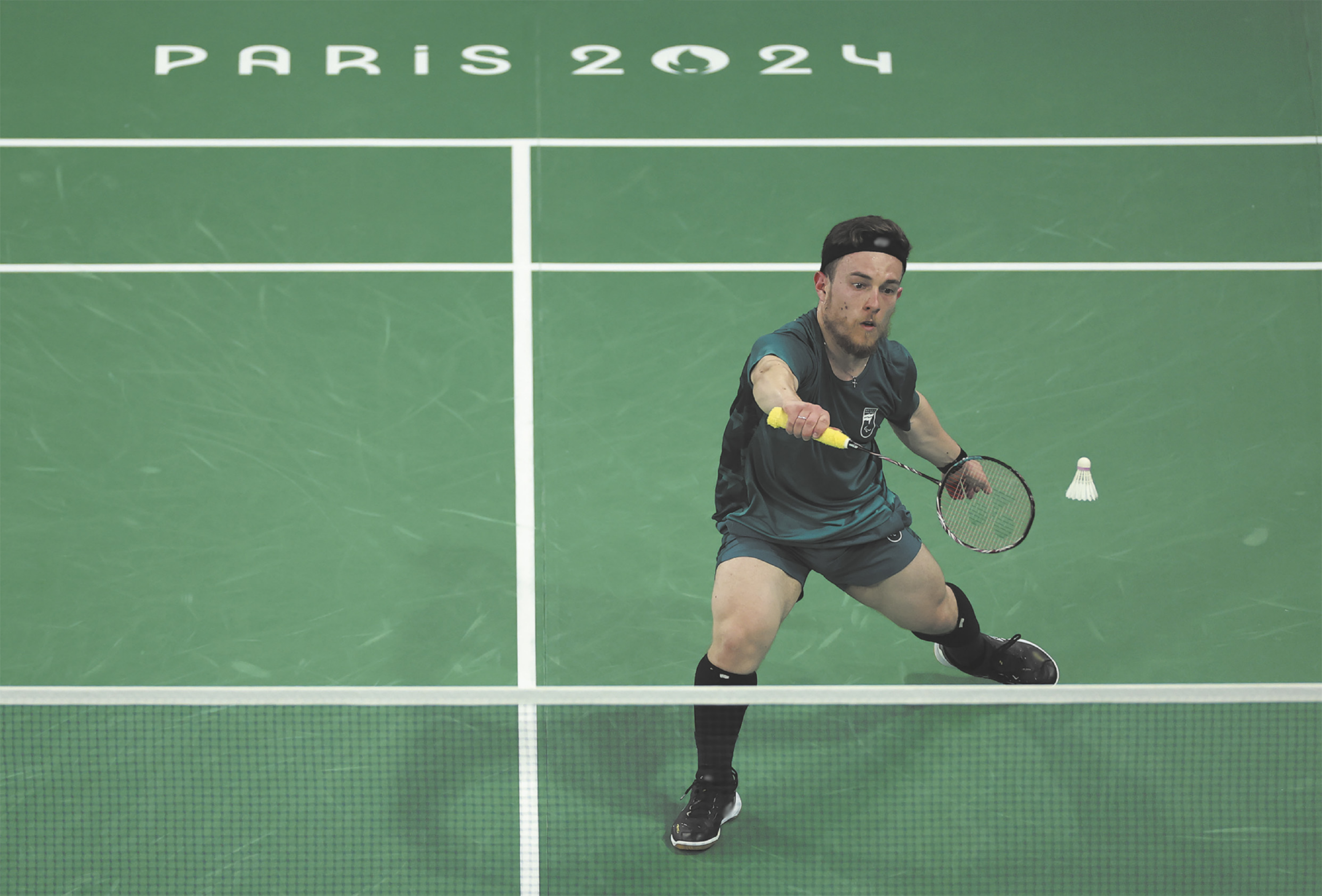Fotografia de Vitor Tavares. Ele é um atleta paralímpico, usando um boné, jogando badminton, com as pernas afastadas e flexionadas, segurando uma raquete diante de uma peteca no ar, ao lado de uma rede.