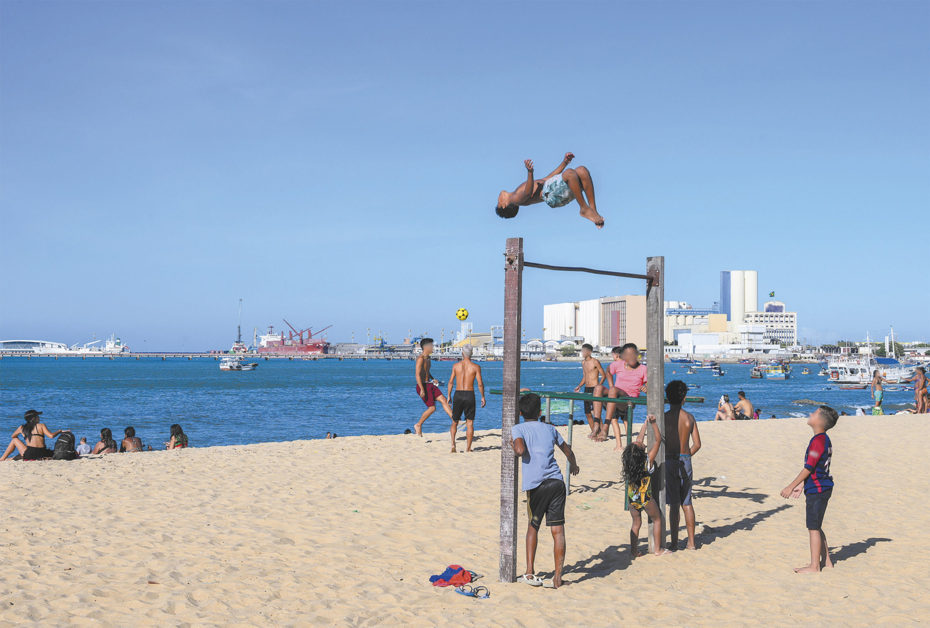 Fotografia de um grupo de crianças reunidas em torno de uma barra de metal horizontal afixada entre dois pilares de madeira, sobre o chão de uma praia. Um garoto está saltando do alto da barra de metal, realizando um salto mortal de costas, em um giro com o corpo deitado no ar, com as pernas flexionadas. Ao fundo há uma paisagem marinha com barcos e edifícios à beira do mar.