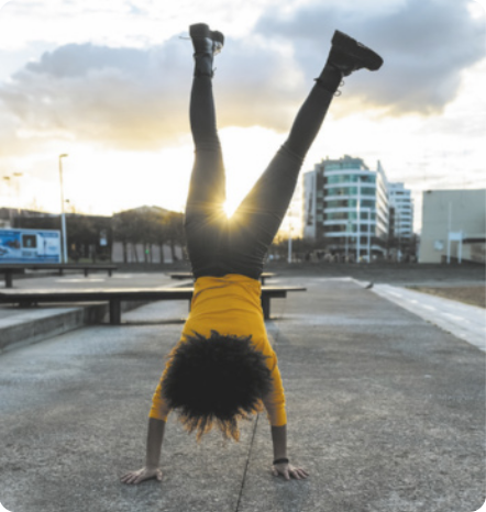 Fotografia de uma mulher de cabeça para baixo sobre um piso de concreto em um espaço público urbano. Ela está com as mãos apoiadas no chão e os braços estendidos, erguendo as pernas estendidas para o alto, se equilibrando com o corpo alinhado.