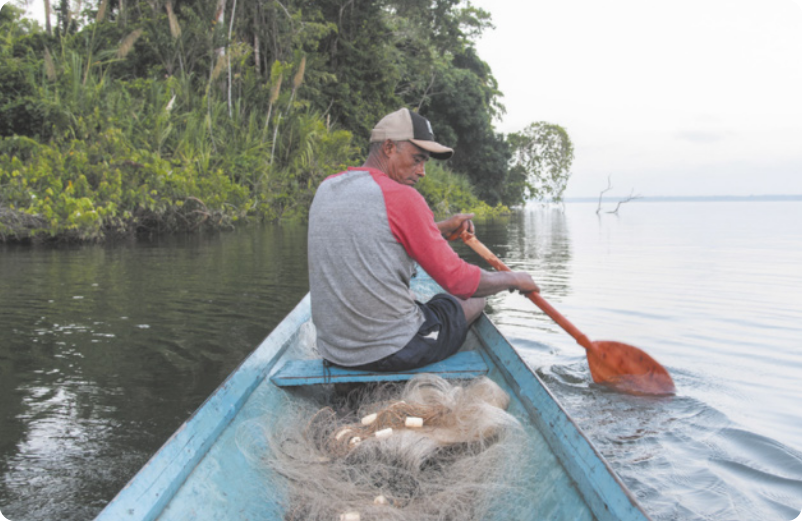 Fotografia de um homem de boné sentado em uma canoa, carregando redes de pesca, remando com um remo curto em um rio largo, com a mata à margem do rio.