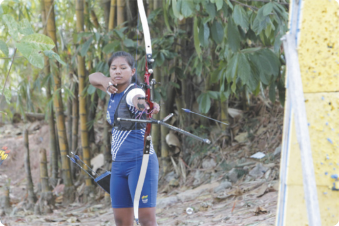 Fotografia de uma garota indígena de uniforme, atirando com um arco e flecha esportivo em direção a um painel com um alvo, com a mata ao fundo.