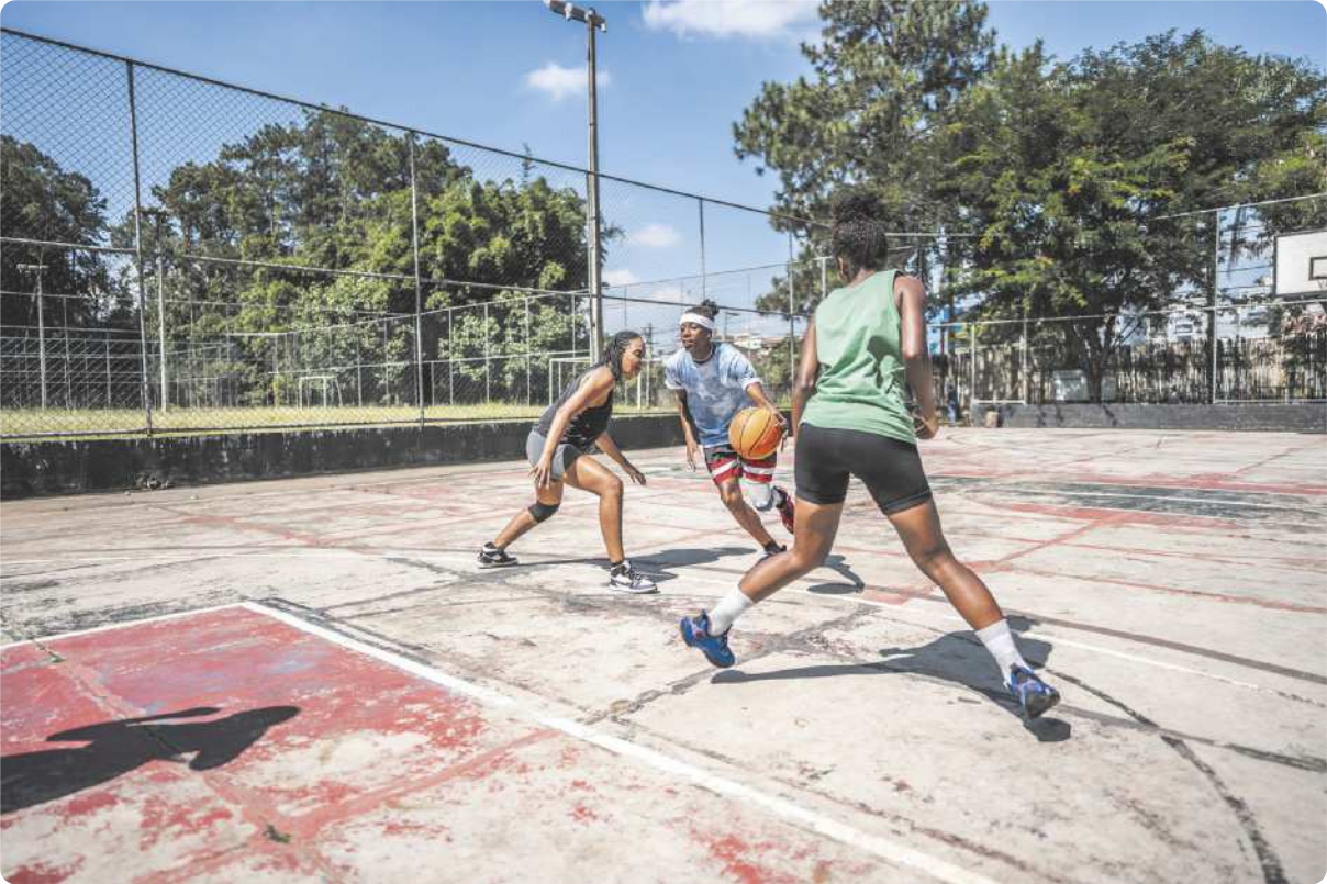 Fotografia de três mulheres jogando basquete em uma quadra pública. Duas delas estão defendendo e uma delas está com a bola, atacando, quicando a bola em direção à cesta.