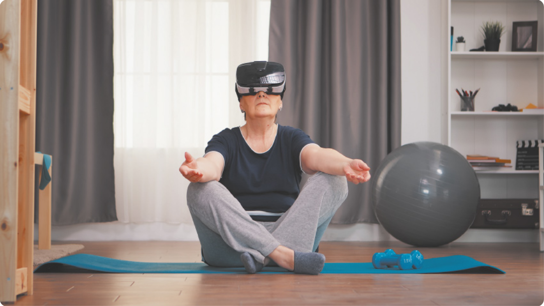 Fotografia de uma mulher sentada em um colchonete estendido no chão, com as pernas cruzadas e os braços estendidos à frente, com os cotovelos apoiados sobre os joelhos, usando óculos de realidade virtual.