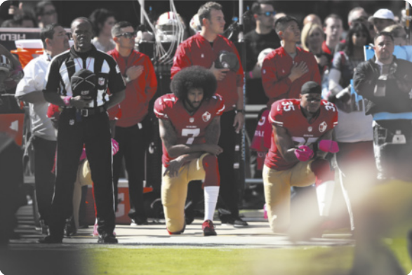 Fotografia de Colin Kaepernick: um atleta negro com barba e cabelos cacheados, vestido com um uniforme de futebol americano, ajoelhado no chão com os braços cruzados apoiados no joelho esquerdo. Ao lado dele está outro atleta negro também ajoelhado, e atrás deles estão diversas pessoas em pé, algumas delas com a mão sobre o coração.