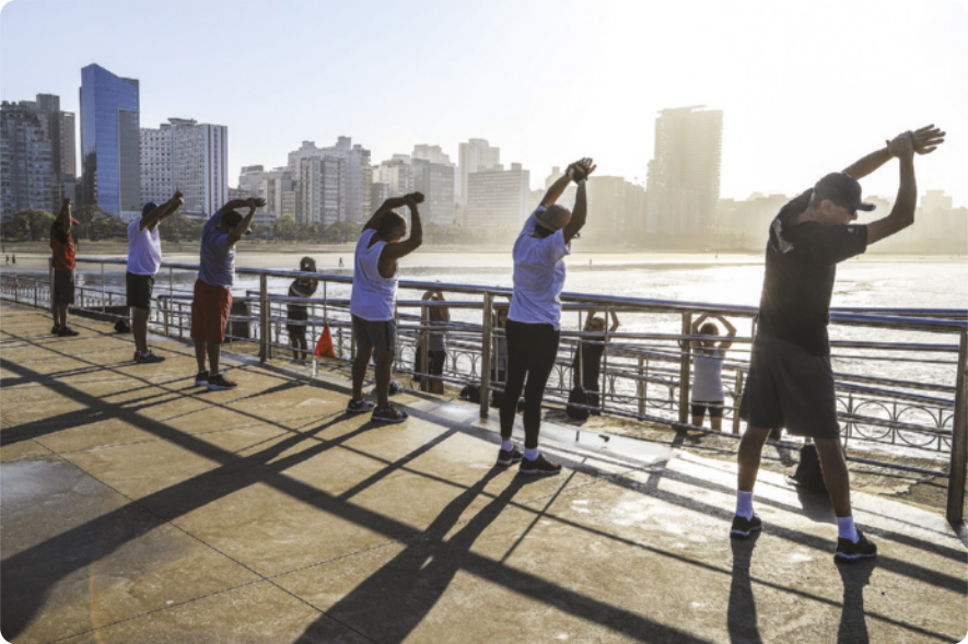 Fotografia de um grupo de homens e mulheres enfileirados à beira de uma praia, em pé, alongando o corpo com os braços erguidos para o alto e inclinando o tronco para a direita, puxando o braço esquerdo com a mão direita.