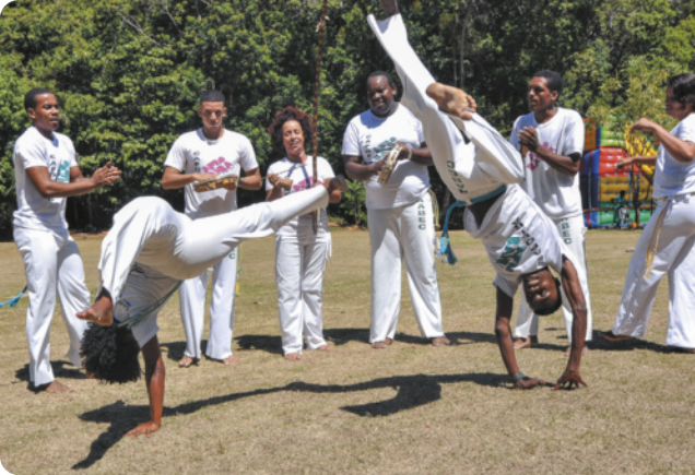 Fotografia de uma roda de capoeira com dois homens jogando ao centro, realizando movimentos acrobáticos com a cabeça para baixo e as pernas afastadas no ar, apoiados com uma das mãos no chão. Em torno deles há pessoas em pé, enfileiradas, batendo palmas, cantando e tocando instrumentos.