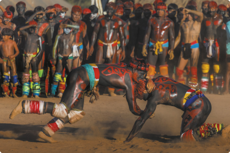 Fotografia de dois homens indígenas usando adereços coloridos e pinturas corporais lutando sobre a areia. Eles estão se agarrando pelos braços, com o corpo próximo ao chão, um deles ajoelhado, e o outro pisando na ponta do pé, caindo em direção ao chão. Ao fundo, há homens indígenas com os mesmos trajes dos lutadores, assistindo a luta em pé.