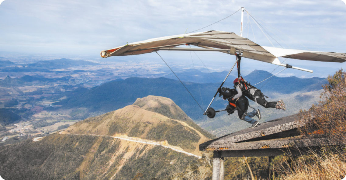 Fotografia de duas pessoas saltando de um telhado no alto de uma paisagem montanhosa, voando em uma asa-delta. Uma pessoa está à frente, saltando e conduzindo a asa-delta, enquanto a outra está atrás dela, deitada no ar, segurando as costas da pessoa à frente.
