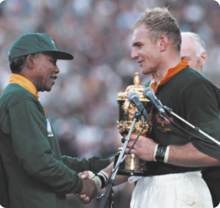 Fotografia de Nelson Mandela. Ele é um homem idoso de boné, cumprimentando com um aperto de mãos o atleta François Pienaar: um homem de cabelos louros curtos, segurando uma taça dourada.