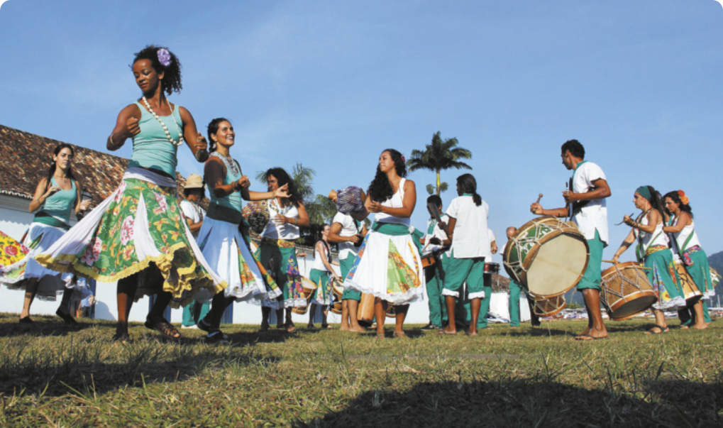 Fotografia de um grupo de mulheres dançando em um gramado, usando saias floridas, colares e flores no cabelo. Diante delas há homens e mulheres tocando tambores.