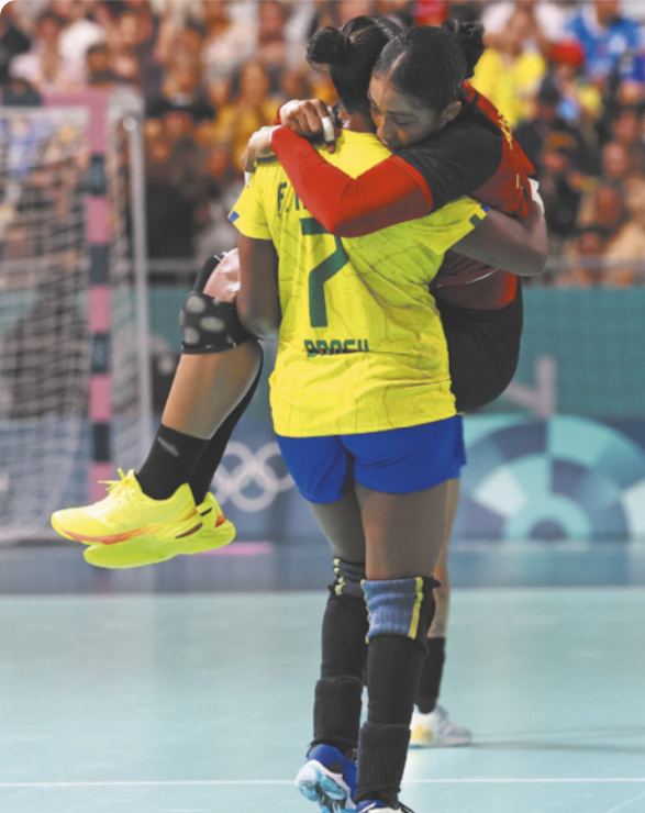 Fotografia de Tamires Araújo, uma mulher com uniforme da seleção brasileira de handebol, carregando no colo a jogadora angolana Albertina Kassoma, que abraça a adversária, apoiando o queixo no ombro de Tamires. Elas estão em uma quadra de handebol e, ao fundo, há uma plateia em uma arquibancada.