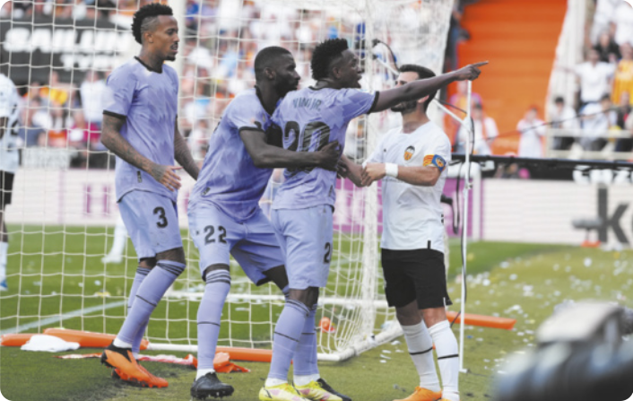 Fotografia de Vinicius Júnior em um campo de futebol estendendo o braço em direção à torcida, com o dedo indicador apontando para a arquibancada. Ele está sendo abraçado e segurado por colegas de time e por um jogador do time adversário.