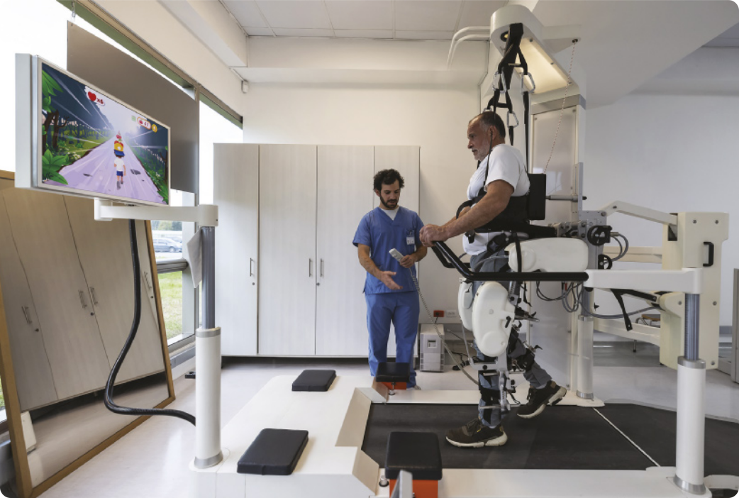 Fotografia de um homem caminhando sobre uma esteira rolante com o auxílio de próteses nas pernas, acopladas a equipamentos mecânicos. Ele está se exercitando diante de uma tela que mostra a imagem de um jogo virtual em que um personagem está correndo em uma estrada. Ao lado dele, um fisioterapeuta acompanha o exercício do paciente.