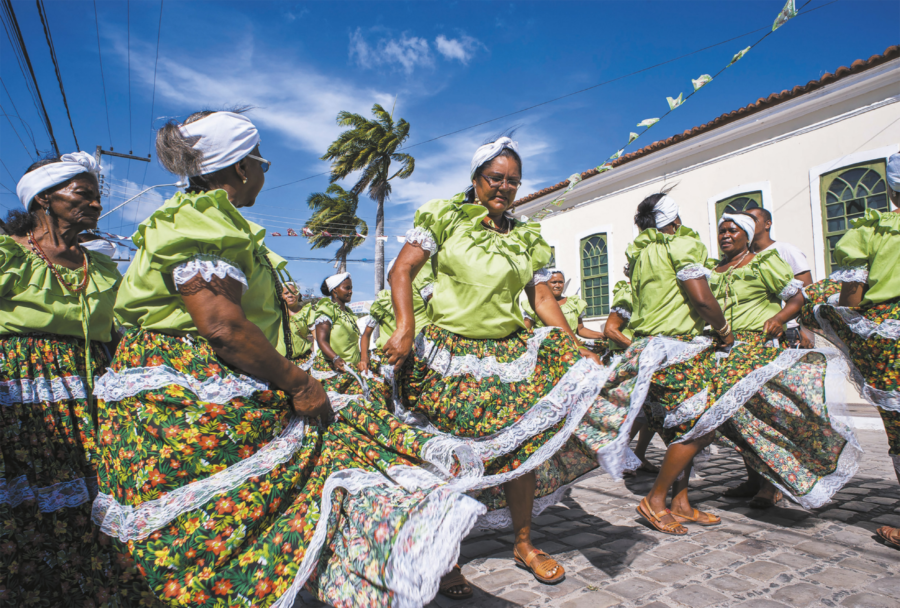 Fotografia de um grupo de mulheres uniformizadas dançando em uma via pública com piso de pedras, usando vestidos com saias floridas e lenços na cabeça.