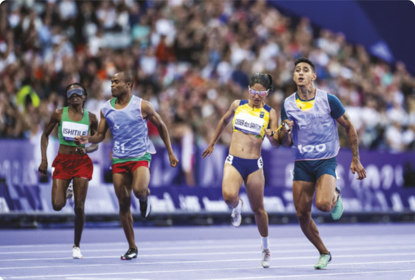 Fotografia de duas mulheres paratletas correndo em uma pista de atletismo, com os olhos vendados, cada uma acompanhada por seu guia. À esquerda, Lahja Ishitile corre ao lado de Sem Shimanda; à direita, Jerusa Geber dos Santos é guiada por Gabriel Aparecido dos Santos Garcia.