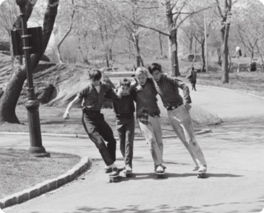 Fotografia em preto e branco de quatro garotos andando de skate lado a lado, abraçados, em uma via asfaltada de um parque arborizado.
