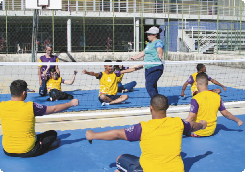 Fotografia de um grupo de pessoas se exercitando em uma quadra de vôlei sentado, com a rede baixa dividindo os dois lados da quadra. Eles estão sentados no chão, com os braços abertos ao lado do corpo, três de cada lado da quadra, acompanhando as instruções de uma treinadora que está em pé, de braços abertos.