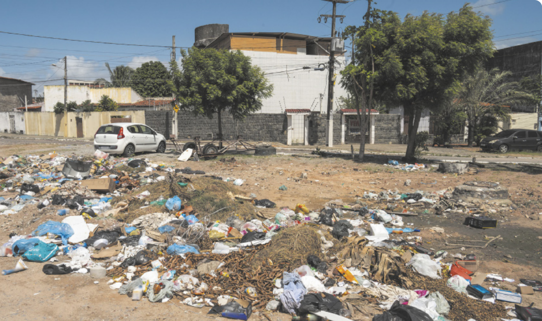 Fotografia de uma praça em que há uma grande pilha de lixo e resíduos diversos no chão, com algumas árvores e casas ao fundo.