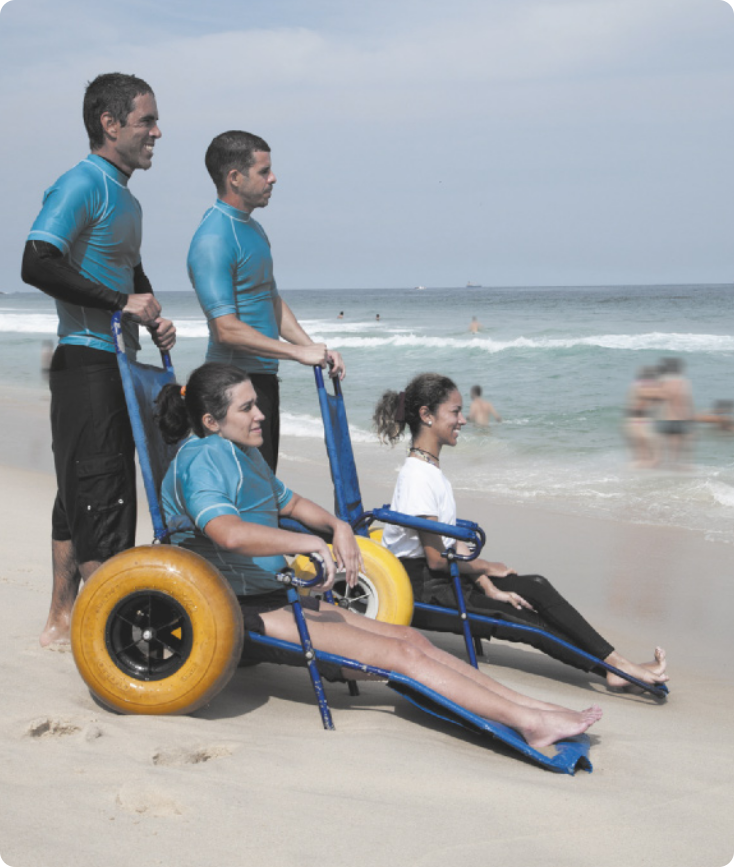 Fotografia de duas mulheres sentadas em cadeiras com rodas de pneus largos, na areia à beira do mar, com dois homens atrás delas, conduzindo as cadeiras.