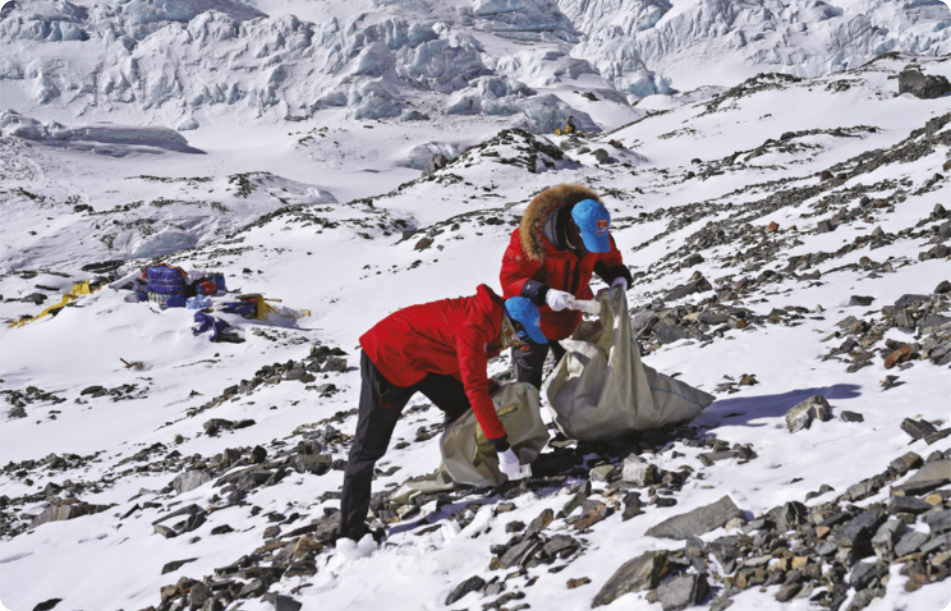 Fotografia de duas pessoas com roupas de inverno segurando sacolas e recolhendo lixo em uma paisagem montanhosa e nevada.