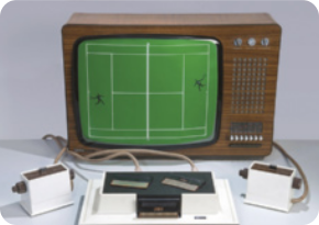 Fotografia de um videogame com o console retangular, com dois controladores de jogo com a forma de caixas com botões. O videogame está conectado a um monitor por cabos, e a tela exibe uma quadra de tênis vista em planta, com dois personagens, um de cada lado da quadra.