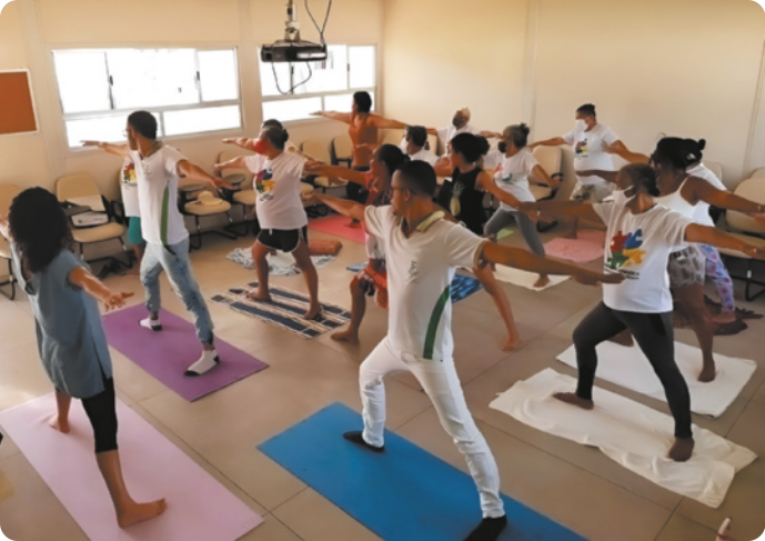 Fotografia de um grupo de pessoas em uma sala, praticando ioga sobre colchonetes estendidos no chão. Uma instrutora está à frente do grupo, em pé, com os braços abertos à altura dos ombros e as pernas afastadas, estendendo a perna direita e flexionando a perna esquerda. As pessoas reproduzem a postura executada pela instrutora.