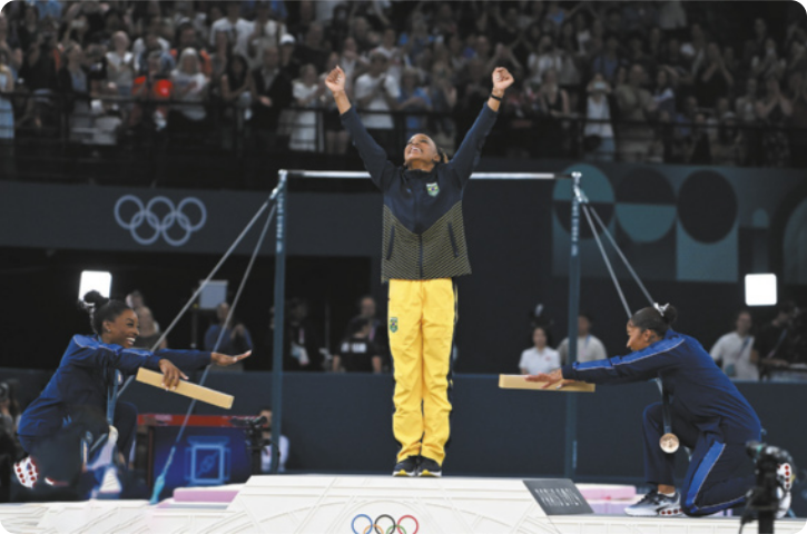 Fotografia de Rebeca Andrade em pé com os braços erguidos para o alto no topo de um pódio, com as atletas Simone Biles e Jordan Chiles ao lado dela, à esquerda e à direita, ajoelhadas diante dela, fazendo gestos de reverência à atleta brasileira. Ao fundo há uma arquibancada com uma plateia e equipamentos de ginástica artística.