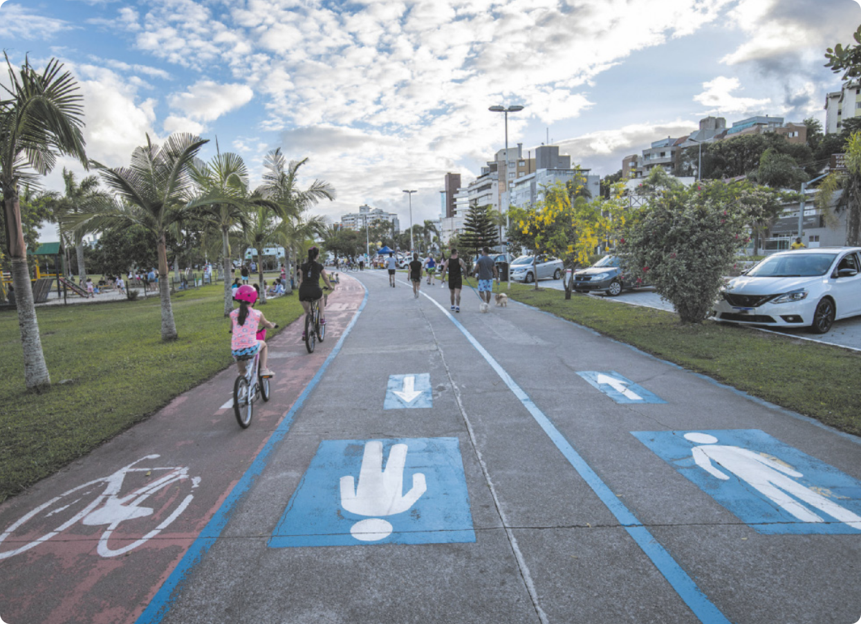 Fotografia de uma pista de asfalto em uma cidade com edifícios baixos, uma praça e canteiros de grama à beira da pista. Pessoas caminham e andam de bicicleta na pista, que é dividida em três faixas: duas reservadas a pedestres, e uma mais estreita, reservada às bicicletas.