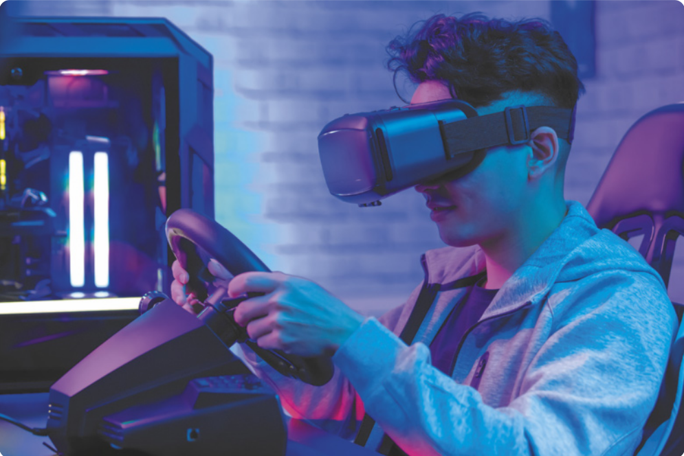 Fotografia de um homem usando um óculos de realidade virtual, sentado diante de um volante para jogos eletrônicos, dirigindo o volante, que está sobre uma mesa ao lado de um computador.