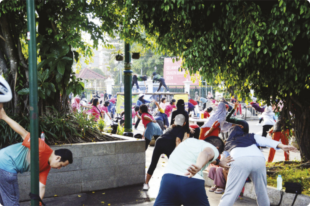 Fotografia de um grupo de pessoas fazendo ginástica em uma praça, com as pernas afastadas e o tronco inclinado para a frente, estendendo um dos braços para a frente, e o outro braço para trás.