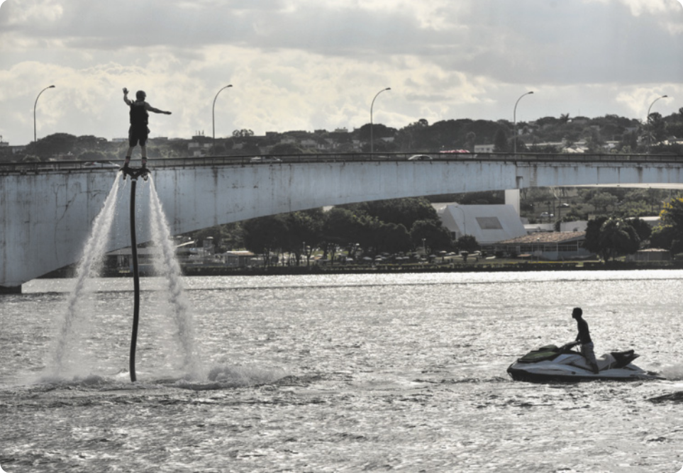 Fotografia de um homem flutuando no ar, sobre a superfície de um lago, com os pés apoiados sobre um flyboard. Para que flutue, o equipamento tem um propulsor que solta jatos de água em direção à superfície do lago, cuja água é puxada por um tubo na base do flyboard que tem a extremidade submersa. Ao lado, está um homem montado em um jet ski, e ao fundo há uma ponte que passa sobre o lago.