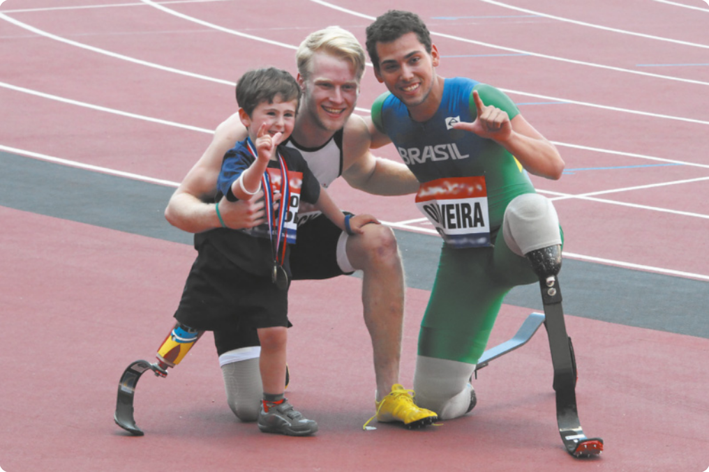 Fotografia de um menino e dois paratletas em uma pista de atletismo. Eles são: Rio Woolf, um menino com uma prótese em uma das pernas, com uma medalha no pescoço; ao lado dele está Jonnie Peacock: um homem com barba e cabelos louros, também usando uma prótese em uma das pernas, ajoelhado, abraçando o menino; e ao lado deles está Alan Fonteles: um homem com próteses nas duas pernas, ajoelhado.