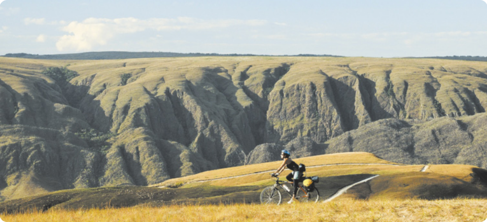 Fotografia de uma mulher andando de bicicleta em uma paisagem montanhosa com vegetação rasteira.