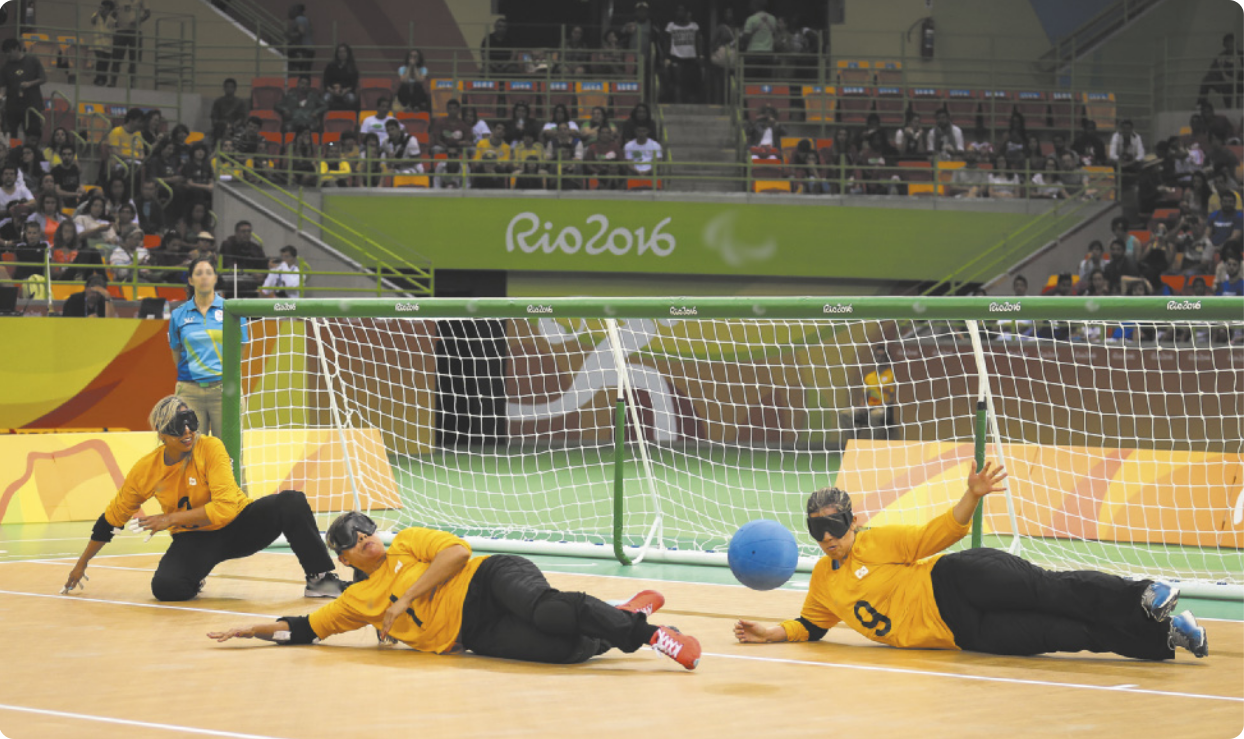 Fotografia de três mulheres jogando goalball, com máscaras sobre os olhos, em uma quadra com um gol largo e baixo ao fundo. Uma delas está ajoelhada no chão e as outras estão deitadas com o corpo de lado, movendo-se em direção a uma bola no ar, que está quicando no chão diante delas.