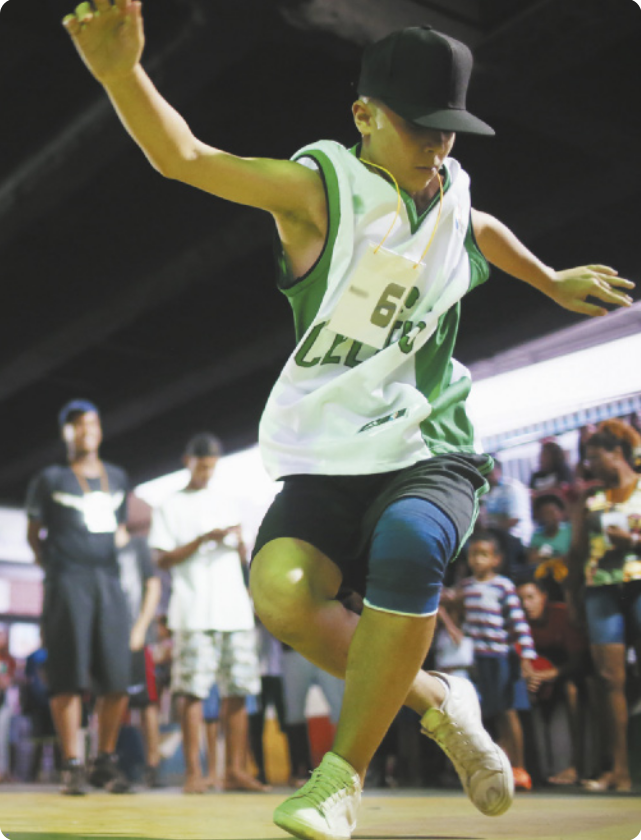 Fotografia de um garoto de boné executando um passo de dança com as pernas cruzadas, uma à frente da outra, flexionando os joelhos e abrindo os braços à altura dos ombros. Ao fundo há um grupo de pessoas em pé assistindo.