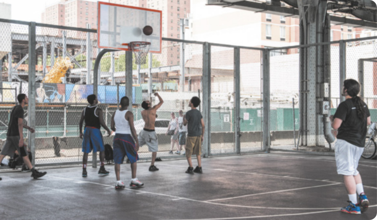 Fotografia de um grupo de homens jogando basquete em uma quadra ao lado de uma via pública, com a calçada ao fundo.