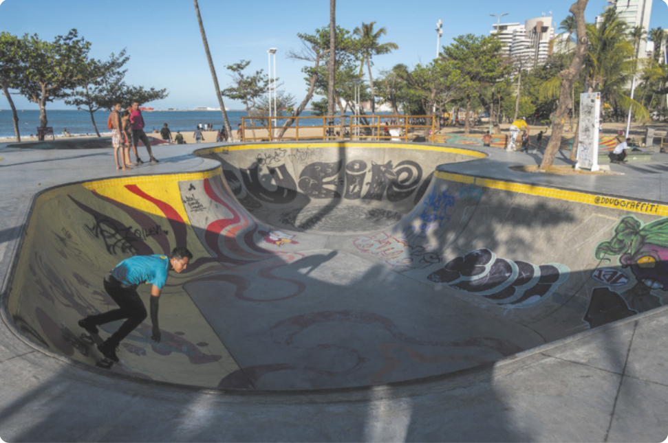 Fotografia de um homem andando de skate em uma pista com formato curvo, semelhante a uma piscina vazia com rampas nas bordas.