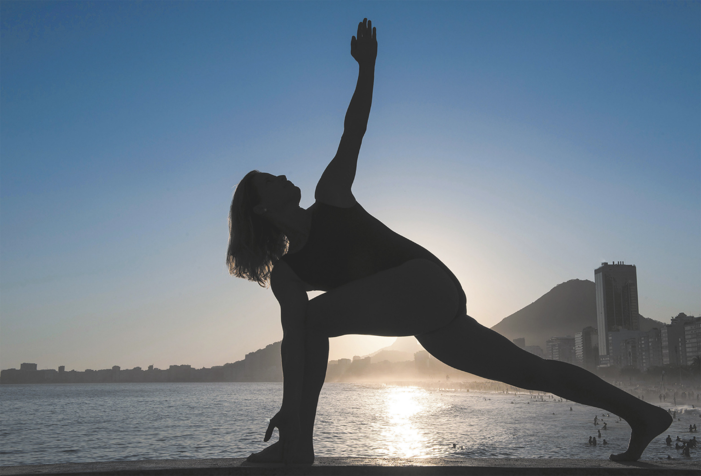 Fotografia de uma mulher de maiô em uma praia, com as pernas afastadas, uma flexionada à frente, e a outra estendida atrás. Ela está com o tronco inclinado para a frente e rotacionado, com um dos braços estendido para o alto, olhando para cima.
