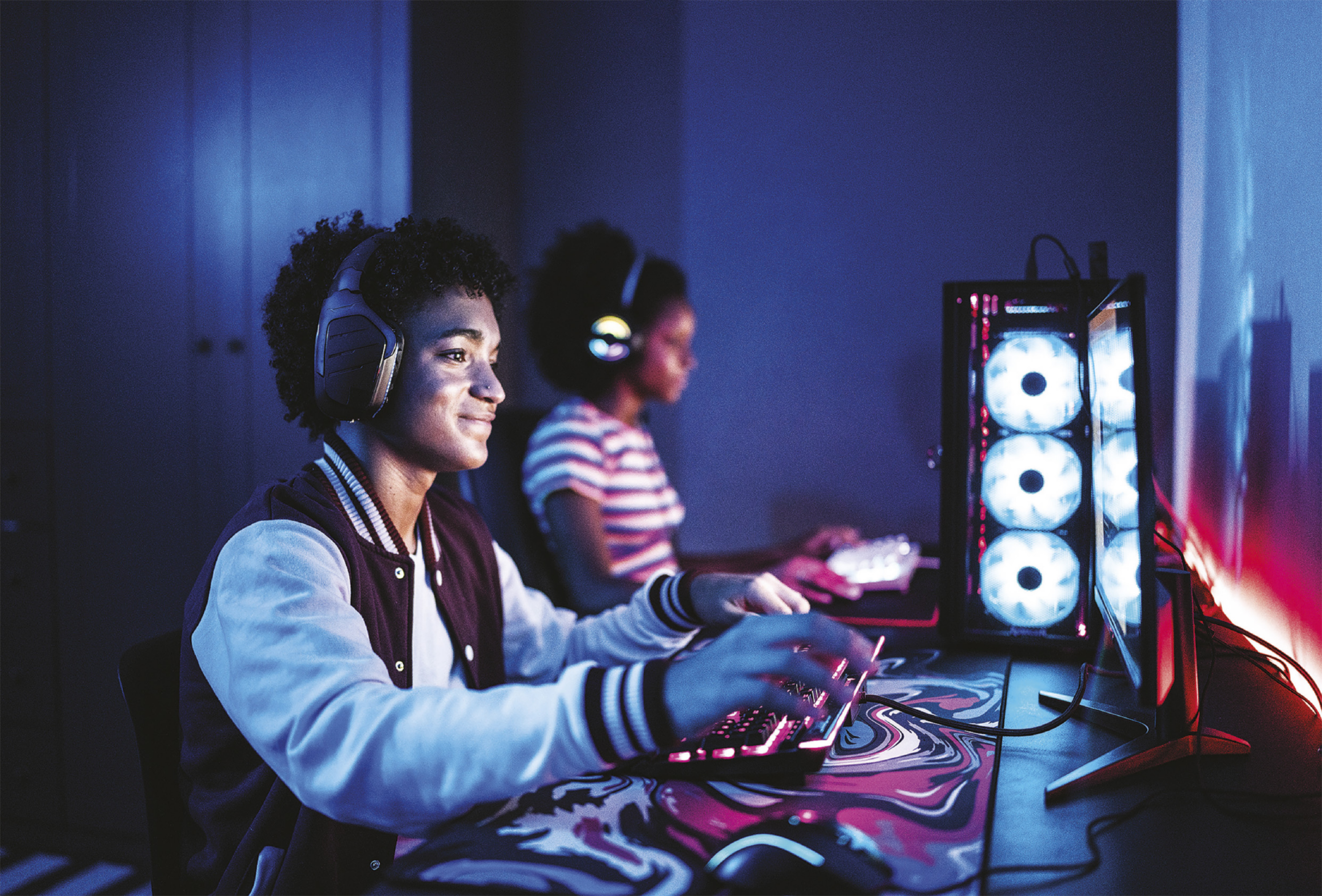 Fotografia de um garoto e uma garota sentados diante de computadores, usando fones de ouvido e manuseando os teclados dos computadores, que têm detalhes luminosos.