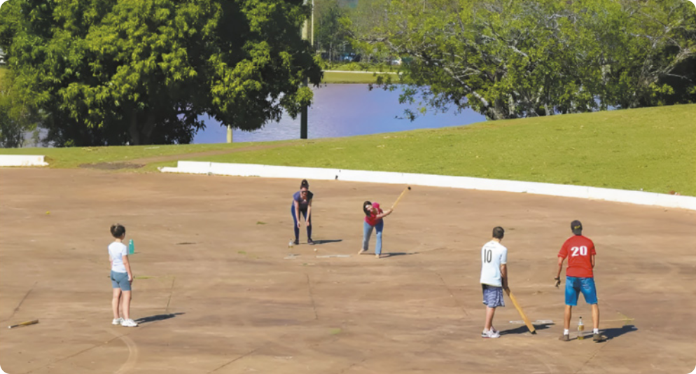 Fotografia de pessoas jogando bets em uma praça com piso de concreto, com um lago ao fundo. Duas garotas estão em uma base, uma delas rebatendo uma bola com um taco e a outra atrás, em posição de defesa; na outra base há dois homens, um deles com um taco na mão e o outro atrás, observando a tacada da rebatedora do outro lado. Ao lado deles, um pouco afastada, está uma garota assistindo à partida.