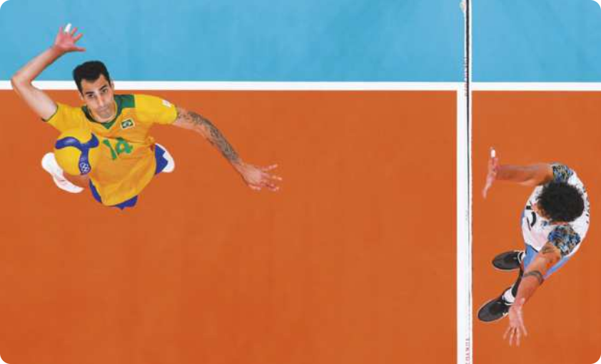 Fotografia de Douglas Souza. Ele é um jogador de vôlei saltando com a mão em direção a uma bola de vôlei, diante da rede, com um jogador adversário do outro lado saltando para bloquear a bola.