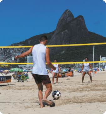 Fotografia de um homem chutando uma bola em uma praia, com uma rede de vôlei diante dele e duas pessoas do outro lado da rede. Ao fundo está o morro do Pão de Açúcar.