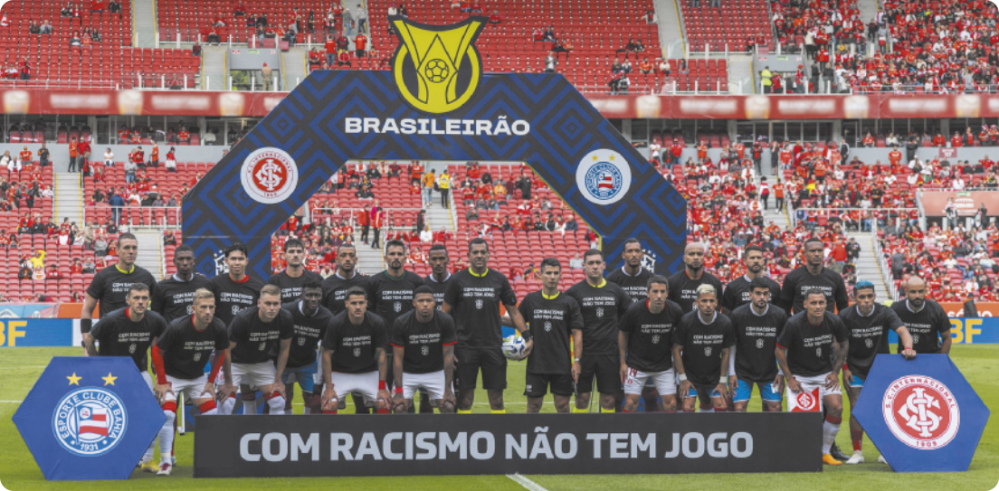 Fotografia dos integrantes de dois times de futebol posando enfileirados em um campo de futebol, todos usando uma mesma camiseta preta, atrás de uma faixa de campanha em que se lê a frase: 'Com racismo não tem jogo'.