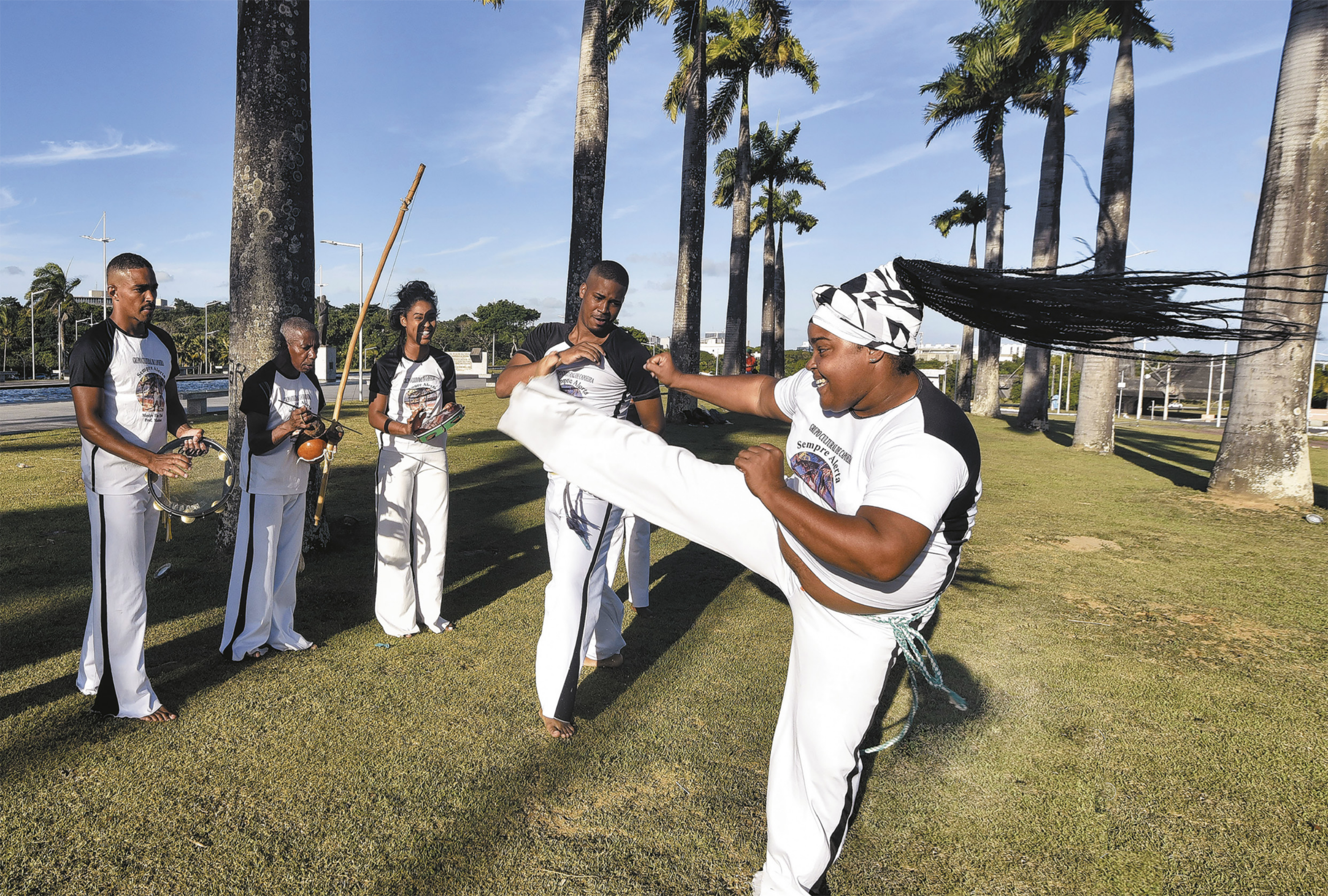 Fotografia de um homem e uma mulher jogando capoeira em um gramado. Ela dá um chute no ar com a perna estendida à altura dos ombros, enquanto o homem ginga diante dela. Em torno deles há uma roda de pessoas em pé cantando e tocando instrumentos como pandeiro e berimbau.