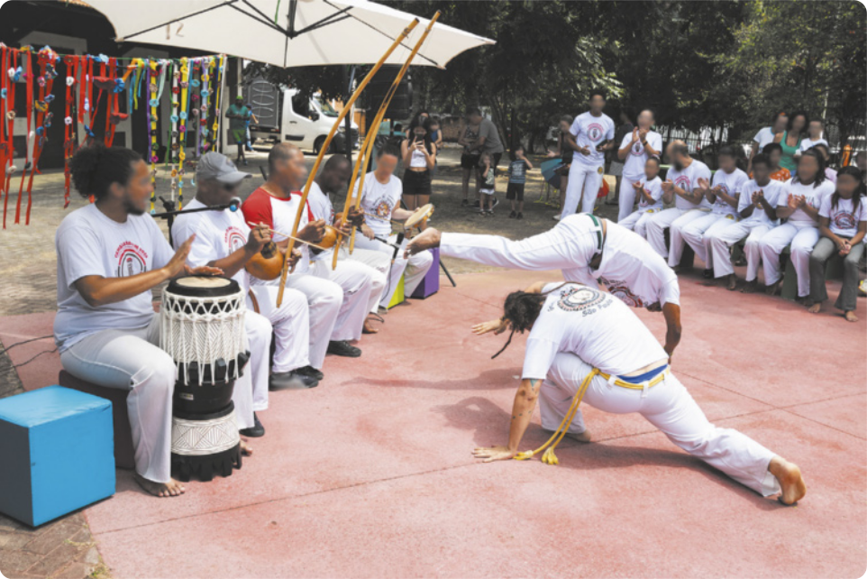 Fotografia de uma roda de capoeira em que dois homens jogam ao centro. Um deles está chutando o ar, com as mãos no chão; e o outro está agachado, se esquivando, com as pernas afastadas e o braço protegendo o rosto. Em volta deles os integrantes da roda estão sentados em bancos, batendo palmas, cantando e tocando instrumentos como berimbau e atabaque.