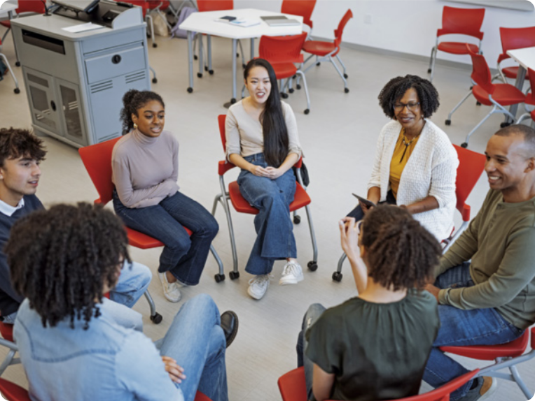 Fotografia de um grupo de pessoas sentadas em cadeiras escolares, formando um círculo. As pessoas são todas jovens, com exceção de uma mulher, que aparenta ser mais velha. Elas estão sorrindo e parecem estar participando de uma discussão em grupo. Ao fundo, há cadeiras, mesas, e um móvel retangular com portas e gavetas.