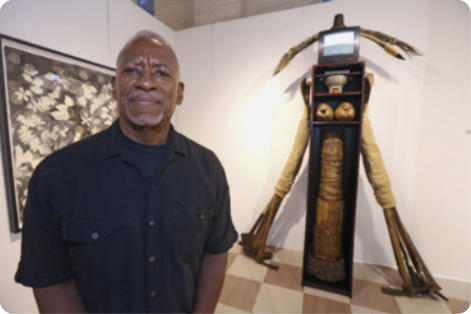 Fotografia de Daniel Lind-Ramos. Ele é negro, tem cabelo e barba grisalhos e está usando uma camisa de mangas curtas, enquanto sorri. Ao seu lado, há uma de suas obras de arte, que se assemelha a uma figura humana. A figura parece ter sido construída a partir de objetos do dia-a-dia da cultura afrocaribenha. Há uma estante alta e estreita de madeira, com uma televisão de tubo, e duas estruturas alongadas dispostas na horizontal, no topo. As prateleiras da estante contém recipientes de cerâmica, o que parecem ser dois cocos, e uma estrutura que se assemelha a um tronco de madeira. Os braços laterais da escultura são feitos de materiais orgânicos, possivelmente fibras naturais ou tecidos. Há um quadro atrás de Daniel.
